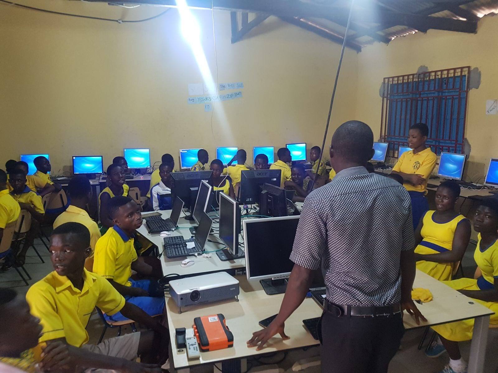 Een computerlokaal op een school in Ghana, met leerlingen in geel-blauwe uniformen achter rijen beeldschermen en een docent vooraan bij de tafel.